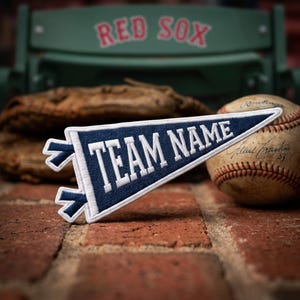 May include: A triangular navy blue felt pennant featuring the white embroidered text TEAM NAME in a bold serif font. The pennant has a white border and two navy blue fabric ties on the left side, presented with a baseball and a glove.