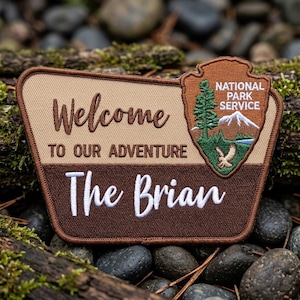 May include: A rectangular embroidered patch with a tan top and dark brown bottom. It features the text Welcome TO OUR ADVENTURE and The Brian in white and brown. A National Park Service shield emblem with a tree, mountain, and bird is attached.