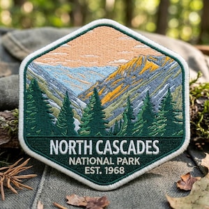 May include: A hexagonal embroidered patch featuring a mountain landscape with dark green pine trees in the foreground, blue and yellow mountain peaks, and a peach-colored sky. The text reads NORTH CASCADES NATIONAL PARK EST. 1968.