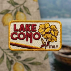 May include: A rectangular embroidered patch with a cream background and a yellow border. The text reads LAKE COMO in bold red letters, with the word ITALY in smaller red text. A cluster of three yellow flowers with brown stems is on the right.