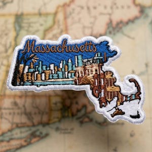May include: An embroidered patch shaped like the state of Massachusetts. The design features a blue sky with the word Massachusetts in brown script, a skyline of buildings in shades of blue and tan, and a white border around the entire edge.