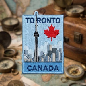 May include: A rectangular embroidered patch featuring a light blue background with the text TO RONTO at the top and CANADA at the bottom in dark blue. The design depicts the CN Tower and a red maple leaf above a grey city skyline.