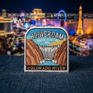 May include: A rectangular embroidered patch featuring a depiction of the Hoover Dam. The design includes brown canyon walls, a white dam structure, and blue water. Text reads HOOVER DAM, BLACK CANYON 1931, and COLORADO RIVER in white and black.