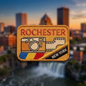 May include: A rectangular embroidered patch with a tan background featuring the red text ROCHESTER at the top. A camera graphic with a film strip is centered above a blue, yellow, and red stripe. A black diagonal corner displays NEW YORK.
