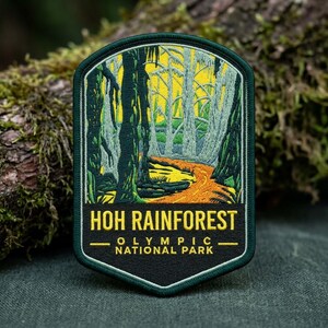 May include: A shield-shaped embroidered patch featuring a forest scene with tall dark green trees, light green mossy branches, and a bright yellow path. The black bottom section displays the yellow text HOH RAINFOREST OLYMPIC NATIONAL PARK.