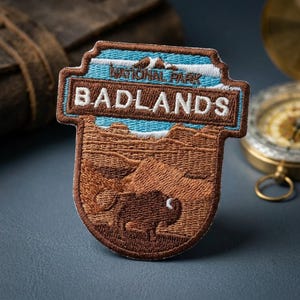 May include: An embroidered patch featuring a landscape of Badlands National Park. The design includes a brown bison in the foreground, layered rock formations, and a blue sky with white text reading NATIONAL PARK BADLANDS in a brown border.