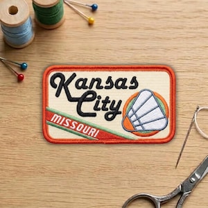May include: Embroidered patch featuring "Kansas City, Missouri" in black script. The patch has a cream background with a red and green stripe and a shuttlecock design. The patch is rectangular with an orange border.
