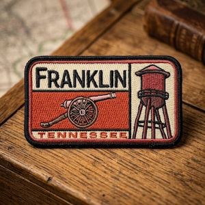 May include: Embroidered patch featuring the word "FRANKLIN" above a cannon and the word "TENNESSEE". The patch also includes a water tower design. The patch has a black border and is set against a wooden surface.