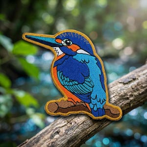 May include: An embroidered patch featuring a kingfisher bird with vibrant blue, orange, and white thread details. The bird is depicted in profile, perched on a brown branch, with a long, pointed beak and a gold-colored stitched border outline.