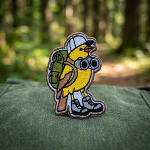 May include: An embroidered patch featuring a yellow bird wearing a white cap, brown hiking boots, and a green backpack. The bird is holding a pair of black binoculars. The patch has a brown stitched border and is shaped like the bird's silhouette.