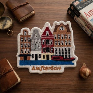 May include: An embroidered patch featuring a row of four colorful canal houses in brown, gray, red, and tan, situated above a blue canal with a red boat. The word Amsterdam is stitched in brown thread at the bottom of the white-bordered patch.