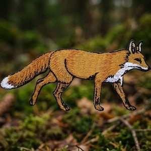May include: An embroidered patch in the shape of a walking fox. The fox features golden-brown fur, a white chest, a white-tipped tail, and black accents on the ears, muzzle, and lower legs, all outlined with a dark border against a blurred background.