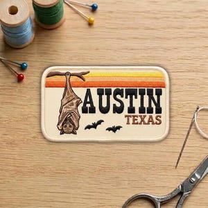 May include: Embroidered patch featuring a bat hanging from a branch, with the text "AUSTIN TEXAS" in black. The background has horizontal stripes in shades of orange and yellow. Two small bats are flying below the text. The patch is rectangular with rounded corners.