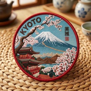 May include: A circular embroidered patch featuring a landscape of Mount Fuji with a snow-capped peak, a traditional temple, and cherry blossom trees. The text KYOTO is stitched in brown, and Japanese characters are present on the right side.