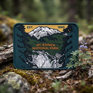 May include: A rectangular embroidered patch featuring a dark teal border and background. The design depicts a snow-capped mountain peak above a forest and waterfall, with the text "EST. 1899" and "MT. RAINIER NATIONAL PARK" in orange.