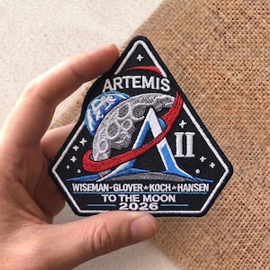 May include: Embroidered Artemis II mission patch. The patch is black, triangular, and features a stylized letter "A" with a blue and red design, a depiction of the moon, and the text "ARTEMIS" and "TO THE MOON 2026".