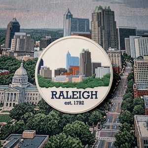 May include: A circular embroidered patch featuring a stylized skyline of Raleigh, North Carolina, with green trees at the base. The text RALEIGH and est. 1792 are stitched in dark blue thread on a cream-colored background with a white border.