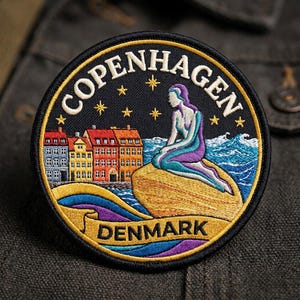May include: A circular embroidered patch featuring the text COPENHAGEN at the top and DENMARK at the bottom. The design depicts a mermaid sitting on a rock, colorful buildings, blue waves, and yellow stars against a black background.