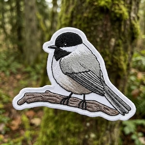 May include: An embroidered patch featuring a black-capped chickadee perched on a brown tree branch. The bird has a black head and throat, white cheeks, and gray and tan wings. The patch has a white border and is shaped to the bird's outline.