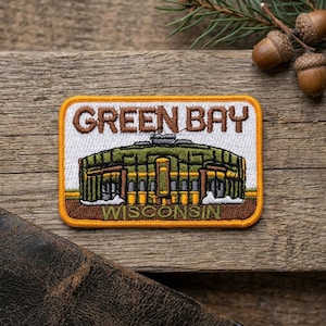 May include: A rectangular embroidered patch featuring the text GREEN BAY in brown letters at the top and WISCONSIN in brown letters at the bottom. The center depicts a stadium in shades of green, yellow, and brown, bordered by a yellow edge.