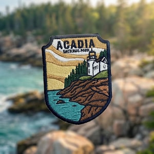 May include: A shield-shaped embroidered patch featuring a landscape of Acadia National Park. The design includes a white lighthouse on a brown rocky cliff, dark green pine trees, a teal blue ocean, and a yellow sky with the text ACADIA NATIONAL PARK.