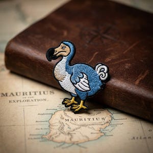 May include: An embroidered patch featuring a dodo bird with blue feathers, a white chest, a tan face, a black beak, and yellow feet. The bird is depicted in profile, standing with its wings tucked against its body and a small curled tail.