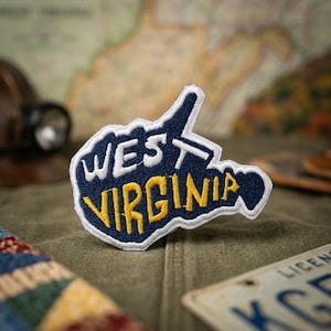 May include: A navy blue embroidered patch shaped like the state of West Virginia with a white border. The word WEST is stitched in white thread, and the word VIRGINIA is stitched in yellow thread across the center of the patch.
