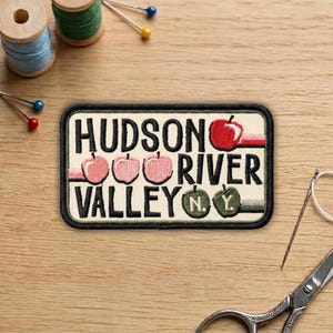 May include: A rectangular patch with the words "HUDSON RIVER VALLEY N.Y." in black lettering. The patch features illustrations of apples in red and pink, with a black border. Sewing supplies, including thread spools and pins, are in the background.