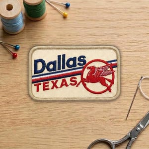 May include: Embroidered patch with the text "Dallas TEXAS" in navy blue and red. A red winged horse is inside a red circle. The patch is rectangular with a tan background and a stitched border.