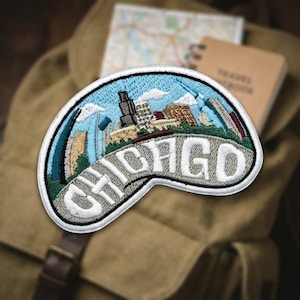 May include: An embroidered patch featuring a stylized Chicago city skyline against a light blue sky with white clouds. The word CHICAGO is stitched in large white capital letters across a grey foreground area, all bordered by a white edge.