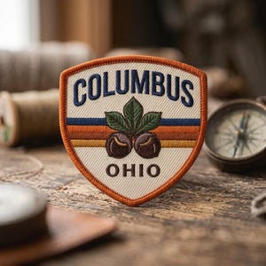 May include: A shield-shaped embroidered patch with an off-white background, featuring the text COLUMBUS in dark blue and OHIO in brown. The center displays two brown buckeye nuts with green leaves above three horizontal stripes in blue, orange, and brown.