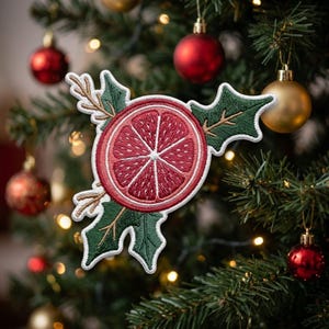 May include: An embroidered patch featuring a central red citrus slice surrounded by dark green holly leaves with light brown stems. The design is outlined in white thread, creating a festive botanical motif with a circular fruit center.