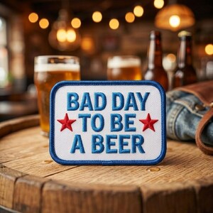May include: A rectangular embroidered patch with a white background and a blue border. The text reads "BAD DAY TO BE A BEER" in blue capital letters, flanked by two small red stars on the left and right sides of the middle line of text.