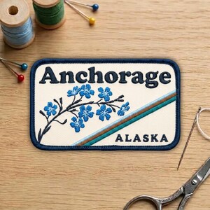 May include: Embroidered patch featuring the word "Anchorage" in navy blue, with a floral design and the word "Alaska". The patch has a navy blue border and a cream-colored background. Sewing supplies are in the background.