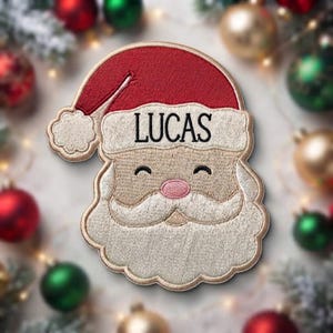 May include: An embroidered patch shaped like a Santa Claus face wearing a red hat with a white pom-pom and trim. The name LUCAS is stitched in black letters on the white hat trim. The face features a pink nose, a white beard, and mustache.