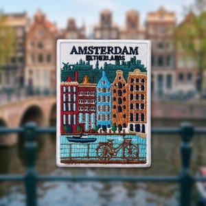 May include: A rectangular embroidered patch featuring a colorful cityscape of Amsterdam, Netherlands. The design includes red, blue, and brown canal houses, a dark blue boat on water, and a brown bicycle. The text AMSTERDAM NETHERLANDS is stitched.