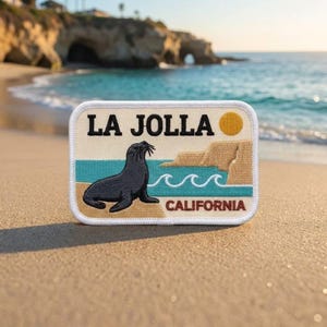 May include: A rectangular embroidered patch featuring a dark gray sea lion on a tan beach, turquoise ocean waves, and tan cliffs. The text reads LA JOLLA CALIFORNIA in black, with a yellow sun circle in the upper right corner, all on a cream background.