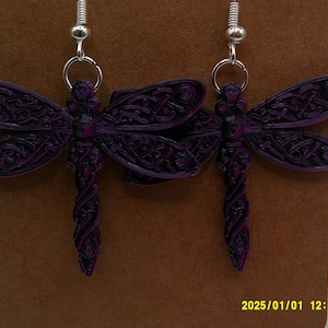 May include: A pair of dragonfly earrings with intricate wing and body details. The earrings are dark purple with black accents, and hang from silver-colored ear wires. The dragonfly design is a Celtic knot pattern.