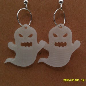 May include: A pair of white ghost-shaped earrings. Each earring features a ghost with an angry face and a wavy bottom. The earrings hang from silver-colored hoops and posts. The background is a brown card.