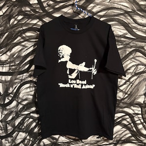 Może przedstawiać: Czarna koszulka z białą sylwetką Lou Reeda z mikrofonem. Tekst "Lou Reed 'Rock n' Roll Animal'" jest wydrukowany poniżej. Koszulka wisi na czarno-białym abstrakcyjnym tle.