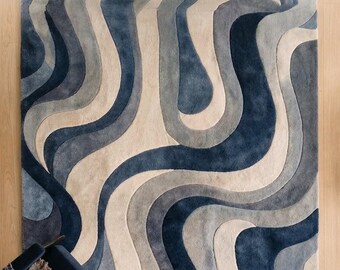 May include: A rectangular area rug featuring a flowing, abstract pattern of undulating waves in shades of navy blue, slate gray, light gray, and cream. The design consists of thick, curved bands that create a sense of movement across the surface.