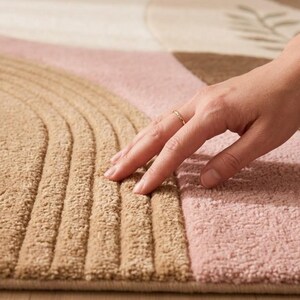 May include: A textured area rug featuring a geometric pattern with raised, carved arch shapes in tan, soft pink, and brown. The rug has a thick, plush pile surface with a visible leaf motif in the corner, showcasing a soft, tufted fabric construction.