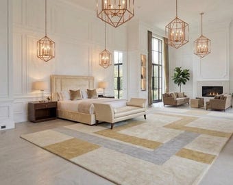 May include: A spacious bedroom featuring a large bed with a beige upholstered headboard, a matching bench at the foot, and a geometric area rug in shades of cream, gold, and gray. Four gold-toned lantern pendant lights hang from the high ceiling.