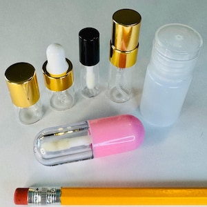 May include: An assortment of small, clear glass bottles and containers with gold, black, and white caps. One container is shaped like a pink and clear capsule. A yellow pencil with a red eraser is in the foreground.