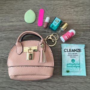 May include: A small pink textured handbag with a gold-tone padlock and tassel keychain. Surrounding items include a light green makeup sponge, a pink nail file, a capsule, a Tic Tac container, a small glass vial, lip balm, and a Cleanze hand wipe.