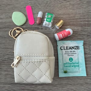 May include: A small, cream-colored backpack keychain with a gold zipper and hardware, next to a green makeup sponge, pink nail file, pills, mints, lip balm, a small perfume bottle, and a packet of antibacterial hand wipes.