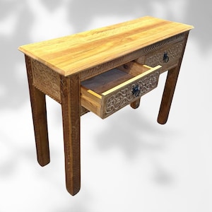 Può includere: Una consolle in legno marrone chiaro con due cassetti, uno aperto. Il tavolo presenta intagli floreali sui frontali e sui lati dei cassetti. Il tavolo è sostenuto da quattro gambe.
