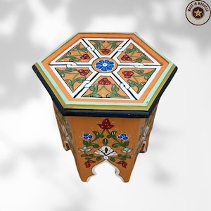 Może przedstawiać: Sześciokątny drewniany stół z ręcznie malowanymi kwiatowymi wzorami w kolorze pomarańczowym, czerwonym, niebieskim i zielonym. Stół ma czarną obwódkę i jasnozieloną lamówkę. Na blacie znajduje się centralny niebieski kwiat i napis "Made in Morocco".