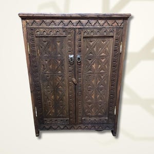 May include: A dark brown wooden cabinet with two doors. The cabinet features intricate carved geometric patterns and decorative trim. The doors have metal ring handles and a latch. The cabinet is a piece of furniture.