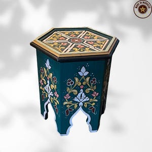 May include: A teal hexagonal side table with floral designs. The top features a geometric pattern with gold, pink, and yellow accents. The sides are adorned with white, pink, and gold floral motifs. The table is a decorative piece.